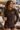 A woman wearing a dark brown long-sleeve rash guard and matching swim shorts stands on a sunny poolside deck holding a drink. The rash guard has 'BON SOUL' printed on the chest in white and pink.
