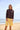 Woman standing on a beach wearing a brown long sleeve rashguard and long yellow and white striped swim shorts. 