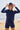 Woman standing on a beach wearing a navy cap with the words LE VIBE in red a navy long-sleeved navy rashguard and navy bather shorts. 