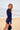 Woman standing on a beach holding a navy cap, wearing a navy long-sleeved navy rashguard top and navy bather shorts. 