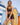 Woman wearing brown bather top and brown bather shorts on beach in front of beach huts holding trainers 