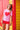 A woman wearing a light pink t-shirt style rashguard with a large red heart graphic and red shorts, leaning against a colorful red and yellow wooden wall.