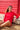 Woman sitting on a sandy beach leaning against a beach hut, wearing a red long-sleeved rashguard and red bather shorts 
