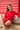 Woman sitting on a sandy beach leaning against a beach hut, wearing a red long-sleeved rashguard and red bather shorts 