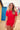 Woman standing on a beach smiling with colourful beach huts in the background, wearing a red short-sleeved rashguard and red bather shorts.