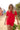 Woman wearing a red short-sleeved rashguard and red bather shorts, walking on a sandy beach with colourful beach huts in the background.