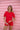 Woman leaning against a pink timber wall, wearing a red short-sleeved rashguard and red bather shorts.