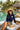 Woman sitting by a pool with palm trees and a hut in the background, wearing a long sleeve navy rashguard featuring a white heart design with the word AMOUR on the left side of her chest
