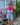 A woman stands outdoors in a backyard garden setting wearing a pink t-shirt with a red heart graphic, light blue denim shorts, olive green sandals, and a green woven shoulder bag.